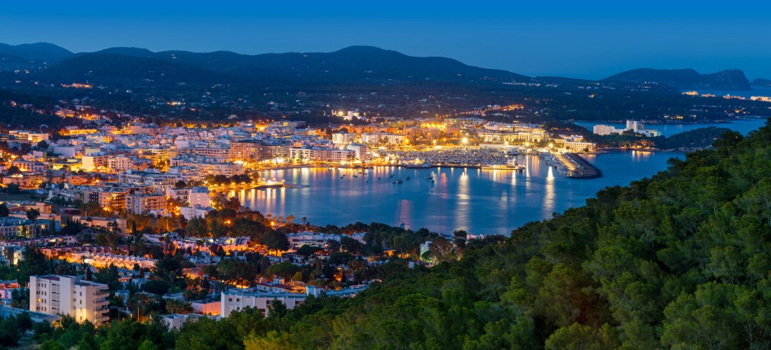Panoramablick auf die Küstenstadt Sant Antoni de Portmany auf Ibiza bei Nacht. Die Lichter der Gebäude spiegeln sich im ruhigen Wasser des Hafens, während bewaldete Hügel im Vordergrund liegen.