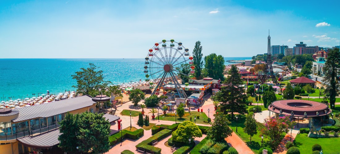 Freizeitpark am Meer mit einem bunten Riesenrad und vielen kleinen Gebäuden. Im Vordergrund gepflegte Grünanlagen, im Hintergrund sieht man zahlreiche Sonnenschirme am Strand und hohe Hotels