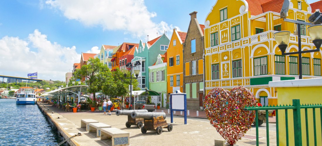 Bunte Häuserfassaden im niederländischen Kolonialstil an der Uferpromenade von Willemstad, Curaçao. Im Vordergrund ein Herz aus Liebesschlössern, alte Kanonen und Sitzbänke am Wasser. Menschen sitzen in Cafés unter schattigen Bäumen, ein Ausflugsboot lieg