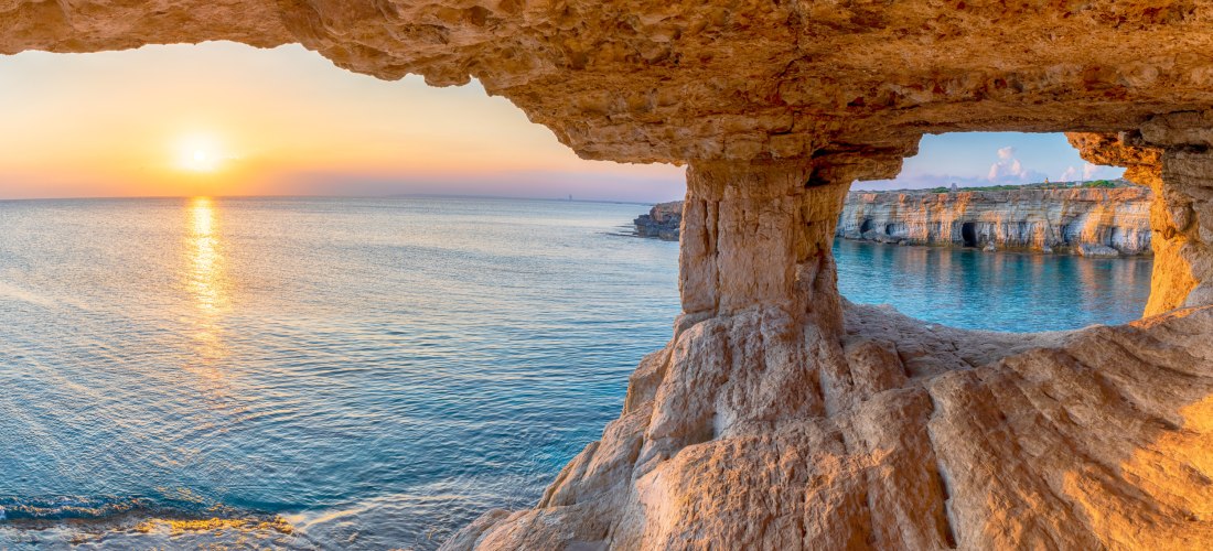 Blick aus einer Felsenhöhle auf das Meer bei Kap Greco auf Zypern mit goldenem Sonnenuntergang und spektakulärer Küstenlinie.
