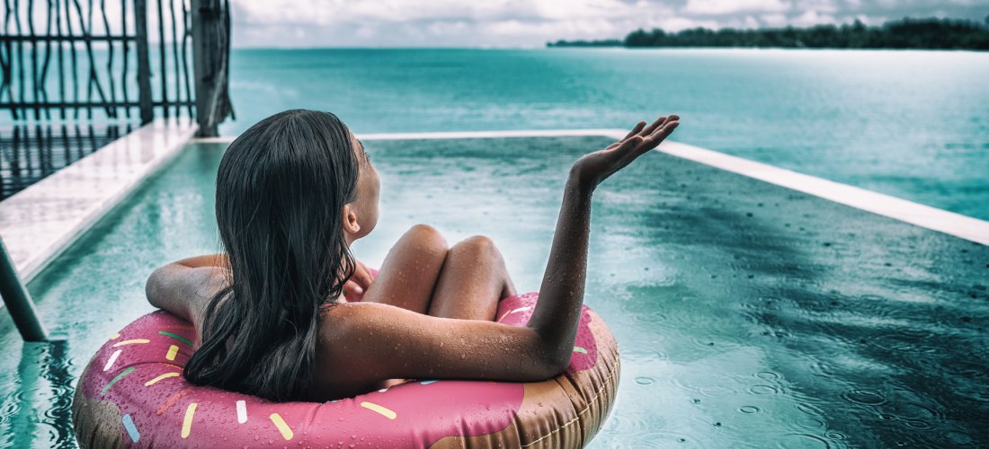 Eine Frau treibt entspannt auf einem Donut-Schwimmring in einem luxuriösen Infinity-Pool mit Blick auf das offene Meer.              Die tropische Kulisse, ein rustikaler Holzsteg und leichte Regentropfen auf der Wasseroberfläche schaffen eine einzigartig
