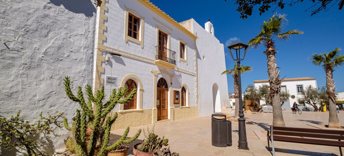 Sonnige Plaza mit weißen Gebäuden im mediterranen Stil in Sant Francesc Xavier, Formentera. Vor dem Hauptgebäude stehen Palmen, ein Kaktus in einem Blumentopf und eine Bank, während eine Person auf einem Fahrrad vorbeifährt.