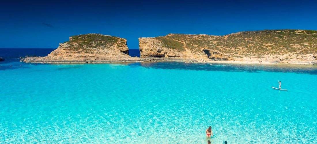 Kristallklares türkisblaues Wasser der Blue Lagoon auf Comino, Malta, mit Badegästen und Paddleboarder unter blauem Himmel