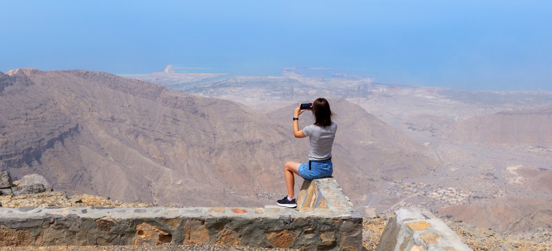 Frau fotografiert die Aussicht vom Jebel Jais auf Berge, Wüste und Meer in Ras Al Khaimah – beliebter Aussichtspunkt im Hadschar-Gebirge.