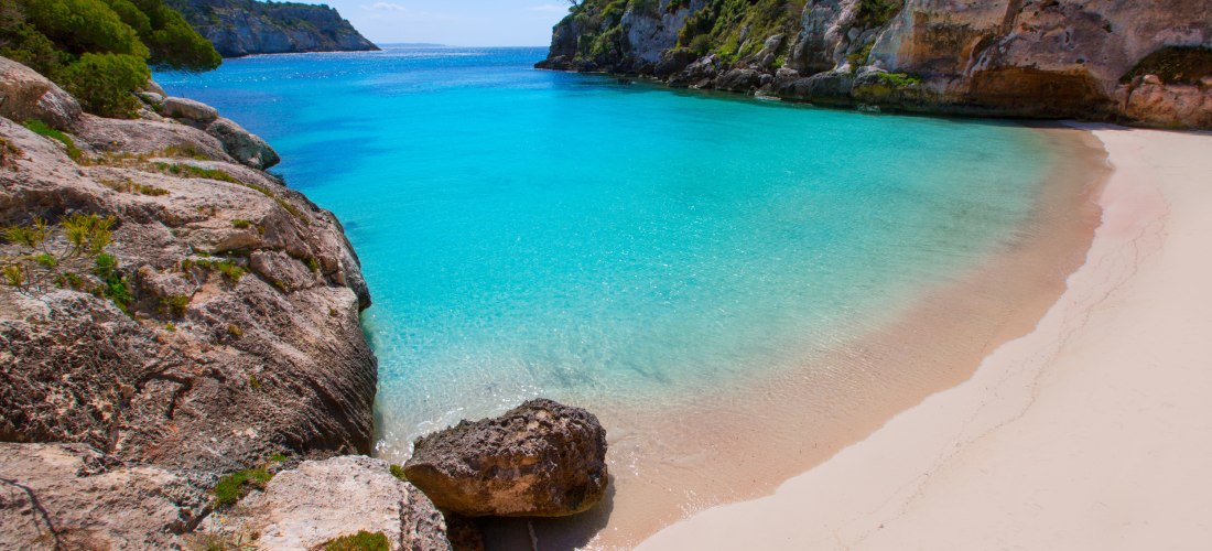 Cala Macarelleta auf Menorca: kleine Badebucht mit kristallklarem Wasser, Felsen und grüner Vegetation. Urlauber schwimmen und entspannen am Strand.
