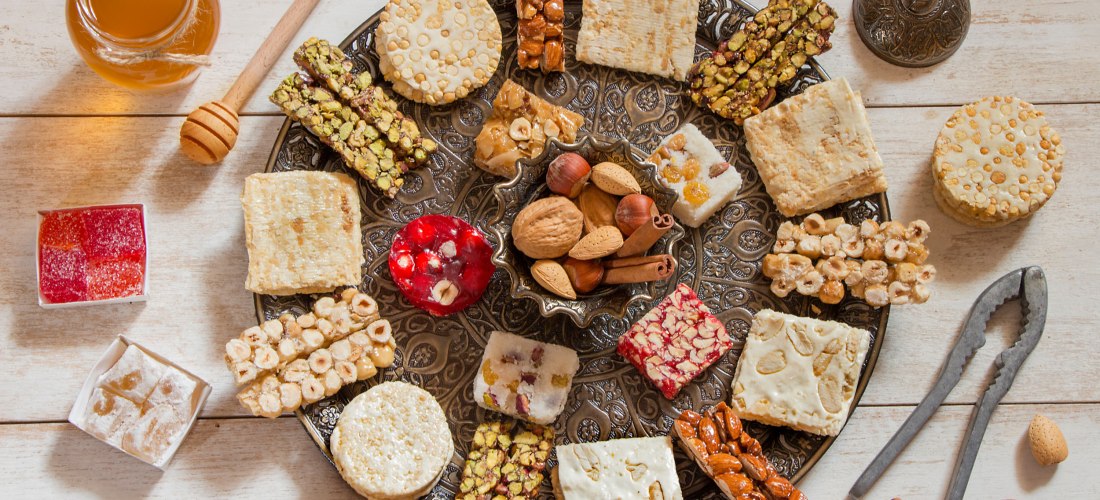 Silberfarbener Servierteller mit orientalischen Süßigkeiten wie Nussriegel, Sesamgebäck, Nougat und türkischem Honig. In der Mitte eine Schale mit Zimtstangen und Nüssen.
