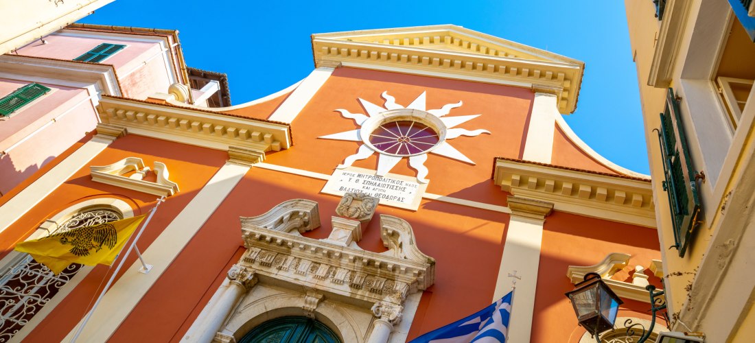 ie Metropolitenkirche von Korfu: Die eindrucksvolle Fassade leuchtet in Orange, flankiert von der griechischen und der byzantinischen Flagge. Über dem Eingang prangt ein weißes Sonnenmotiv – ein Beispiel für die einzigartige Verbindung von Glaube und Arch