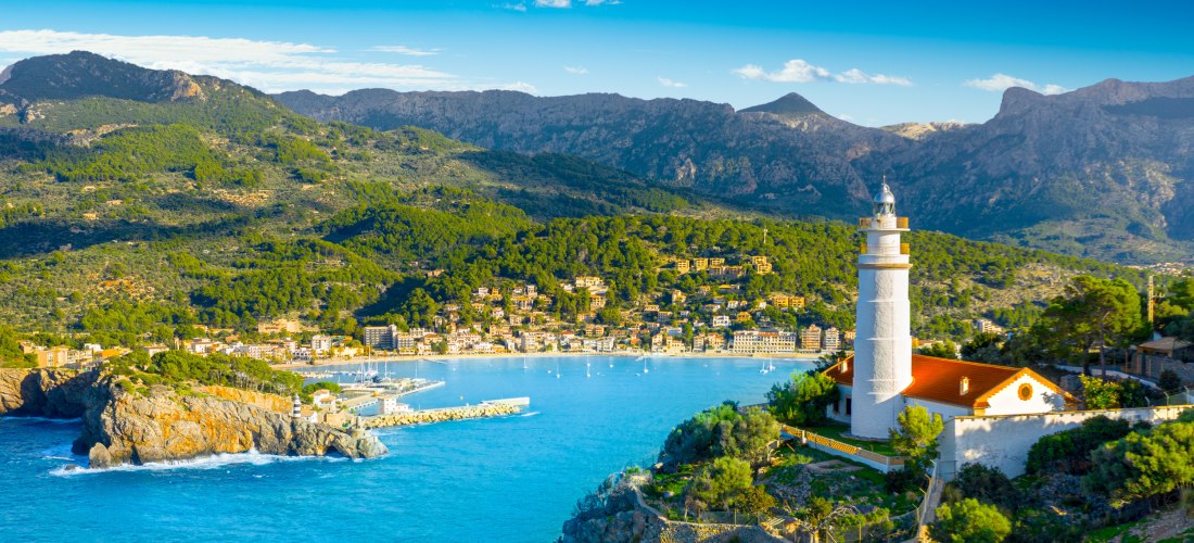Eine wunderschöne Hafenbucht auf Mallorca mit klarem türkisfarbenem Wasser. Segelboote und Yachten liegen im Hafenbecken, während die umliegenden Gebäude und die grüne Landschaft im Hintergrund zu sehen sind. Im Vordergrund ist ein Leuchtturm zu erkennen.