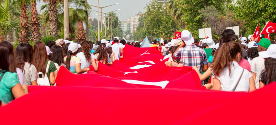 Eine große Menschenmenge, überwiegend junge Menschen, trägt eine riesige türkische Flagge bei einem Umzug durch eine palmengesäumte Straße in einer Stadt.