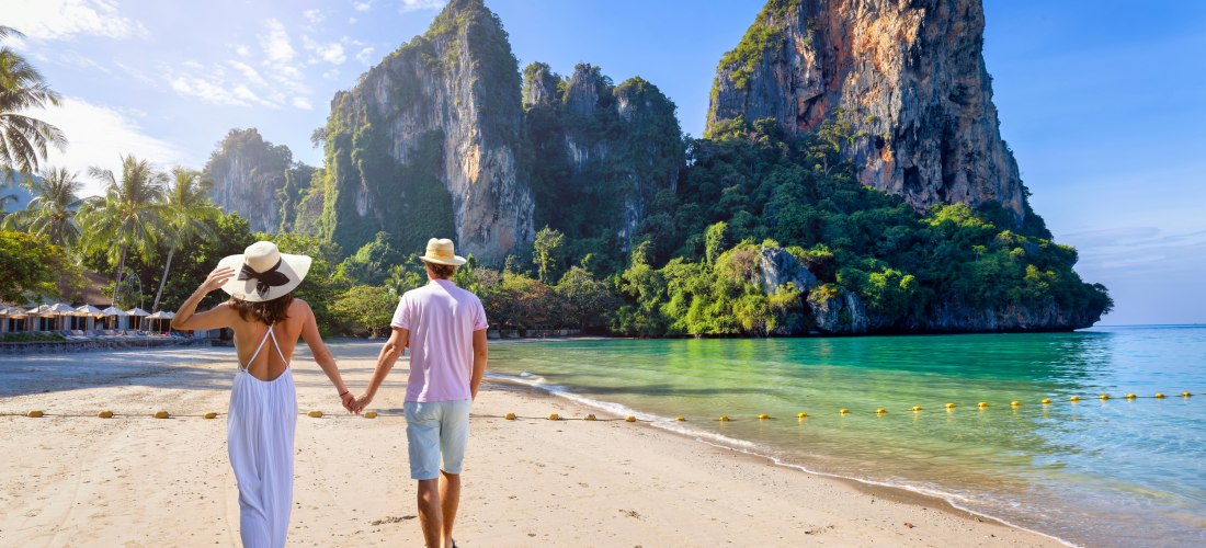 Ein Paar geht Hand in Hand am tropischen Strand entlang. Im Hintergrund ragen grüne Kalksteinfelsen in den Himmel, das Meer schimmert türkis, Palmen säumen den Strand. Die Szene wirkt friedlich und romantisch. ( Railay Beach in Krabi )