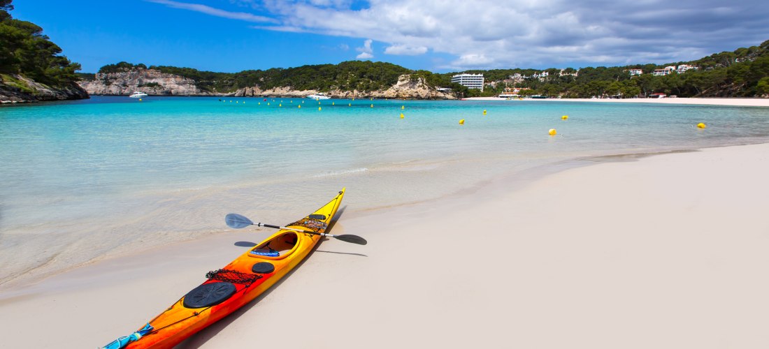Ein rot-gelbes Kajak liegt am weißen Sandstrand einer türkisfarbenen Bucht auf Menorca. Das Wasser ist klar und ruhig, im Hintergrund erstrecken sich bewaldete Felsen und ein Hotelkomplex. Gelbe Bojen markieren den Badebereich. Der Himmel ist teilweise be