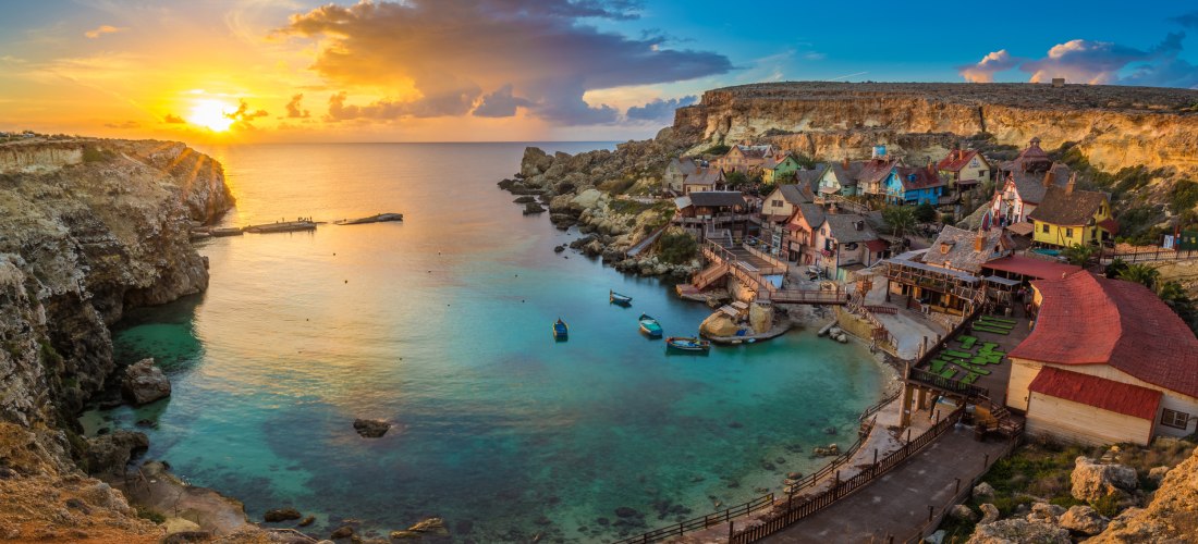 Popeye Village in der Anchor Bay auf Malta bei goldenem Sonnenuntergang, umgeben von türkisblauem Meer und Felsenküste