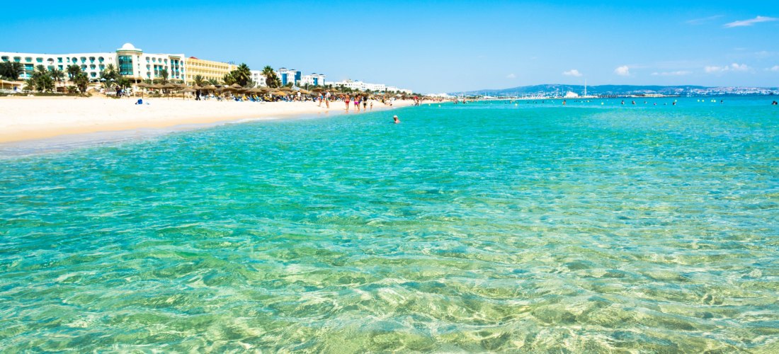 Strand, türkisblaues Wasser, im Hintergrund Hotelanlage, blauer Himmer und Sonnenschein