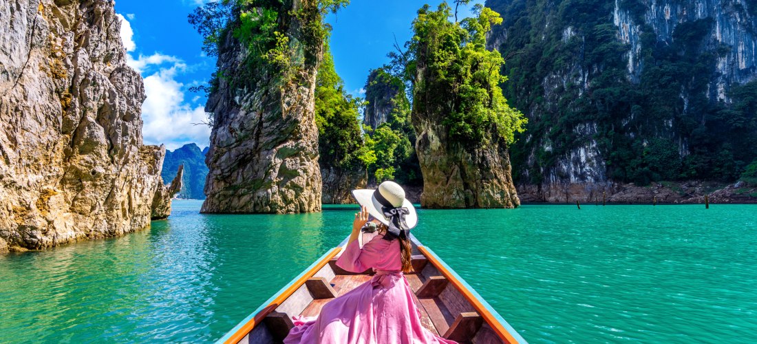 Eine Frau in einem langen rosa Kleid und mit großem Sonnenhut sitzt auf dem Bug eines traditionellen Longtail-Boots. Sie fährt durch türkisfarbenes Wasser inmitten imposanter, steil aufragender Kalksteinfelsen, die mit üppiger tropischer Vegetation bewach