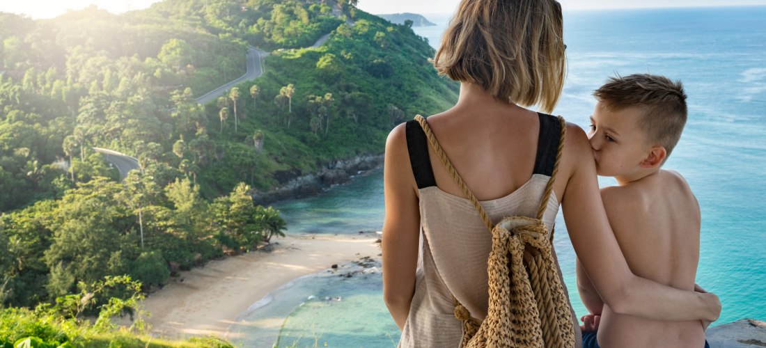 Eine Frau mit heller Sommerkleidung und einem geflochtenen Rucksack sitzt mit einem Jungen, vermutlich ihrem Sohn, auf einer Mauer und blickt auf eine grüne tropische Küste mit einem kleinen, abgelegenen Sandstrand hinab. Der Junge lehnt sich an sie, währ