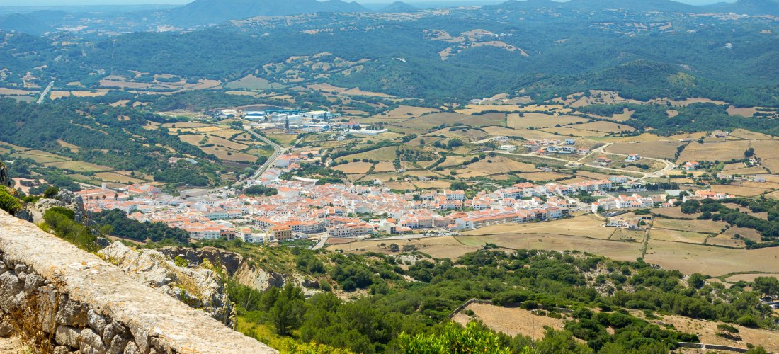 Panoramablick vom Monte Toro auf das Dorf Es Mercadal mit weißen Häusern und roten Dächern, umgeben von Feldern und grünen Hügeln.
