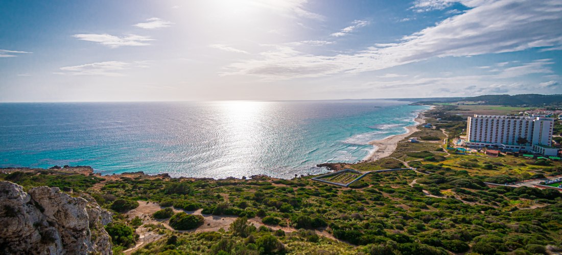 Panoramaaufnahme der Küste von Son Bou auf Menorca mit weiten grünen Dünen, dem langen Sandstrand und einem großen Hotelgebäude am Meer.