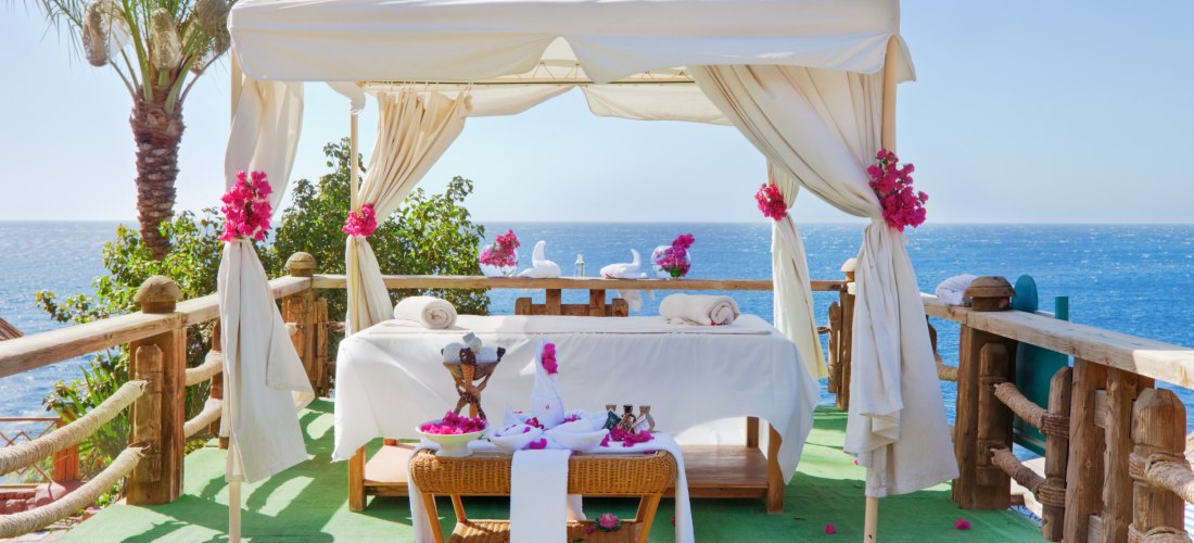 Romantisch dekorierte Massageliege unter einem weißen Pavillon mit Vorhängen und pinken Blüten. Der Platz liegt auf einer Holzplattform mit Blick auf das blaue Meer, umgeben von Palmen und Pflanzen.