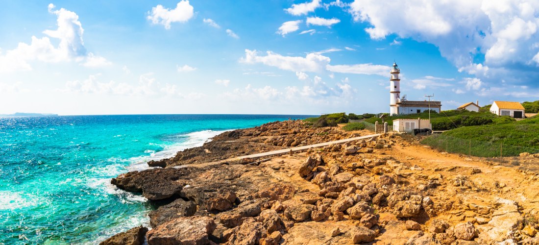 Eine felsige Küste Mallorcas mit schroffen Klippen und türkisblauem Wasser. Auf einem der Felsen steht ein weißer Leuchtturm mit einem roten Dach, flankiert von kleinen Gebäuden. Im Hintergrund wächst spärliche, typisch mediterrane Vegetation. Die Küstenl