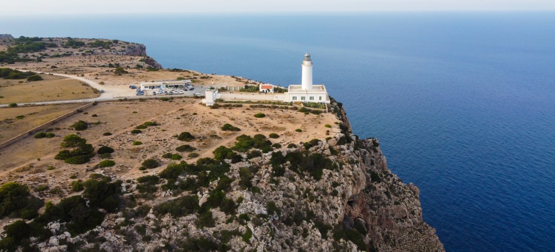 Luftaufnahme des Leuchtturms Far de la Mola auf Formentera. Das weiße Gebäude steht auf einer kargen, felsigen Steilküste hoch über dem tiefblauen Mittelmeer. Parkplätze und kleine Nebengebäude liegen in der Nähe des Leuchtturms.