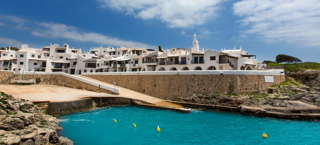 Blick auf Binibeca Vell von der Wasserseite: weiße Häuser mit dunklen Balkonen und Bögen, eine Steinmauer und türkisblaues Meer im Vordergrund.