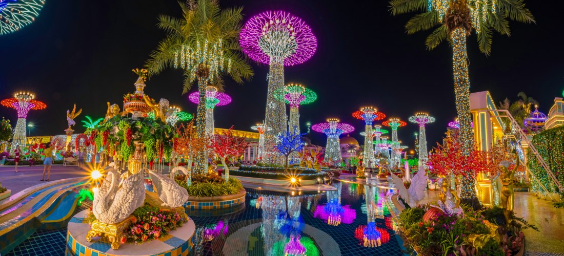 Ein prächtig beleuchteter Themenpark in Pattaya bei Nacht. Riesige, pilzförmige Lichtinstallationen in bunten Farben erleuchten den Himmel. Im Vordergrund ein Brunnen mit Schwanenfiguren und eine spiegelnde Wasserfläche.