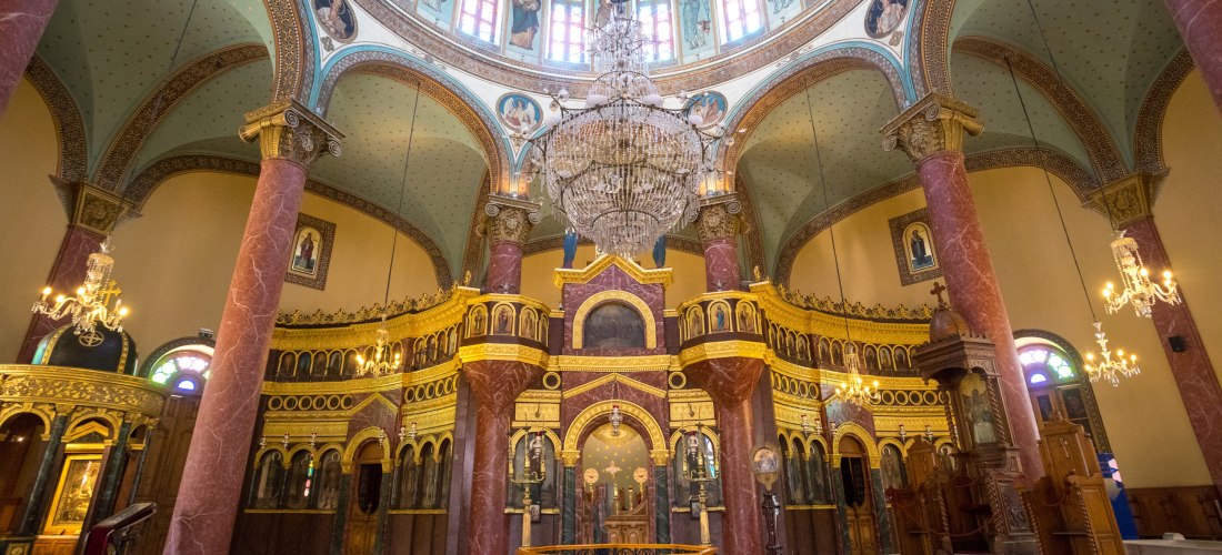 Innenraum einer koptisch-orthodoxen Kirche mit prachtvoller Ikonostase aus Gold und rotem Marmor. Die Wände sind reich verziert, ein großer Kristallleuchter hängt von der zentralen Kuppel, die mit Gemälden von Heiligen geschmückt ist. Hohe Bögen und Säule