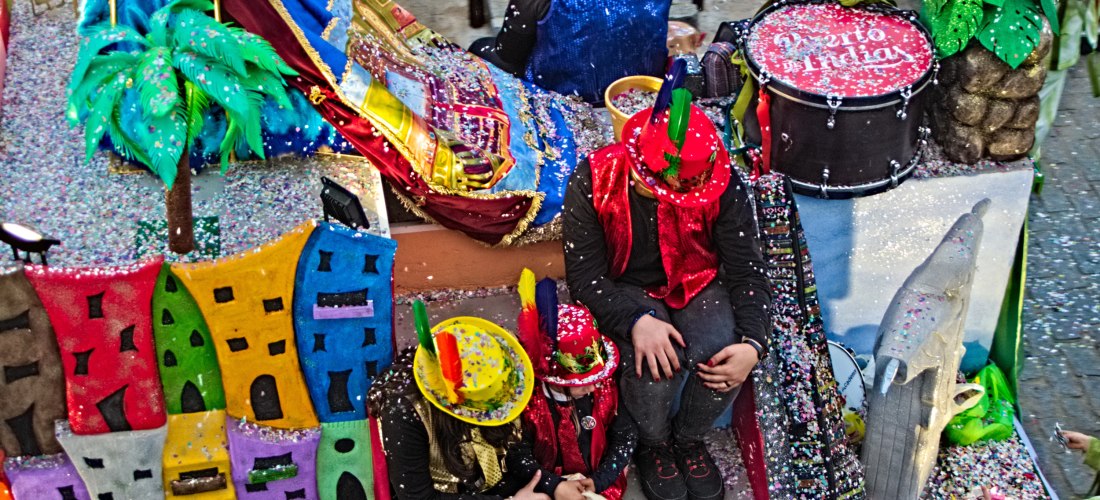 Ein Karnevalswagen mit bunten Dekorationen, Konfetti und fantasievollen Elementen wie einer Palme, einer Stadtfassade und einem Trommelset. Menschen mit glitzernden Hüten und Kostümen sitzen auf dem Wagen. Die gesamte Szene ist von Konfetti bedeckt und st
