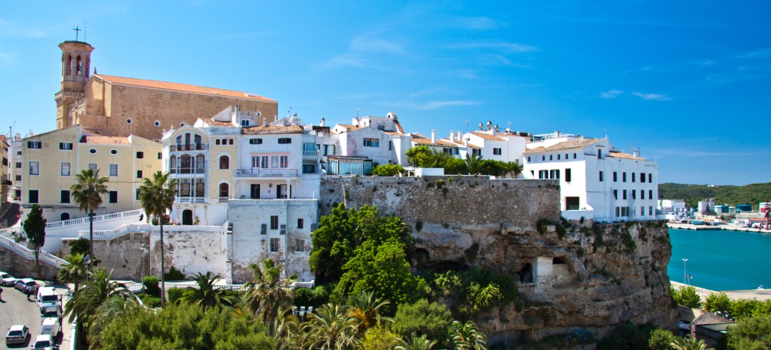 Altstadt von Mahón auf Menorca mit historischen Gebäuden und einer Kirche auf einem Felsplateau. Im Vordergrund Palmen und Blick auf den Naturhafen.