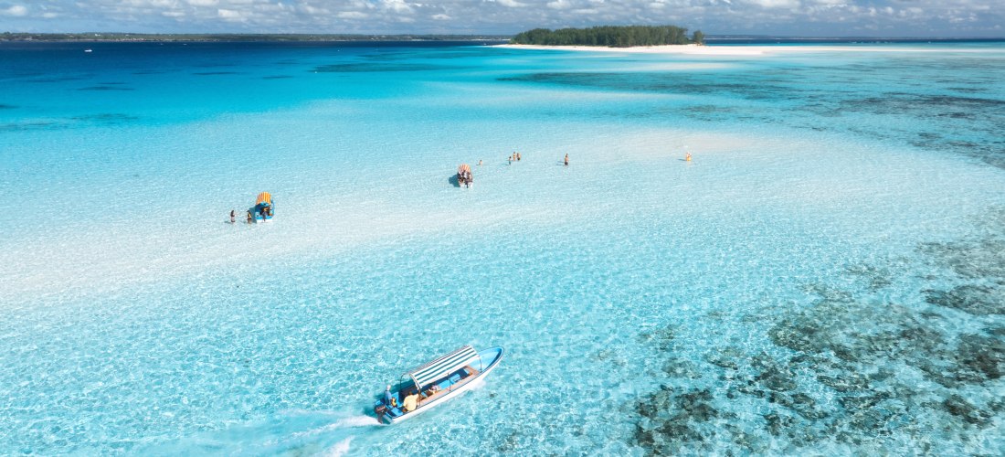 Türkisblaues, flaches Wasser an einer Sandbank vor Sansibar; Badende im seichten Meer und ein kleines Boot mit Sonnendach.