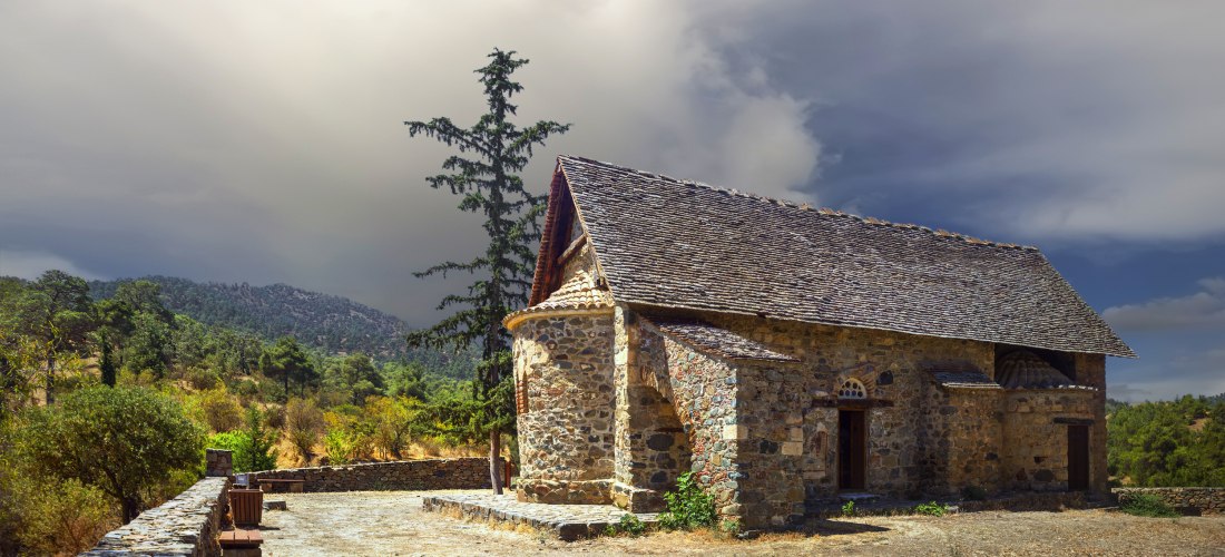 Historische Scheunendachkirche im Troodos-Gebirge auf Zypern: Natursteinbau mit Schieferdach, Zypresse daneben, Berge und dramatischer Himmel.