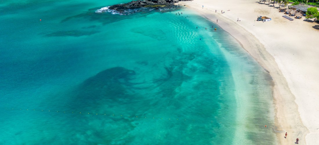 Tropische Badebucht mit türkisblauem Wasser und hellem Sandstrand auf den Kapverden. Am Strand liegen einzelne Badegäste, ein kleines Boot mit Personen treibt im klaren Wasser. Im Hintergrund dichter, grüner Wald und Palmen.