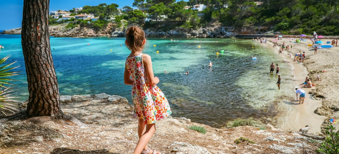 Mädchen in einem bunten Sommerkleid steht auf einer Klippe und blickt auf eine Badebucht mit türkisfarbenem Wasser und Sandstrand auf Mallorca