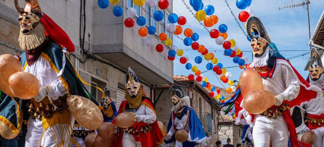 Straßenszene mit traditionellem Karnevalsumzug. Männer in bunten, folkloristischen Kostümen und kunstvollen Masken mit Hörnern und Bärten marschieren durch eine enge Gasse, über der zahlreiche bunte Luftballons aufgehängt sind. Die Teilnehmer halten große