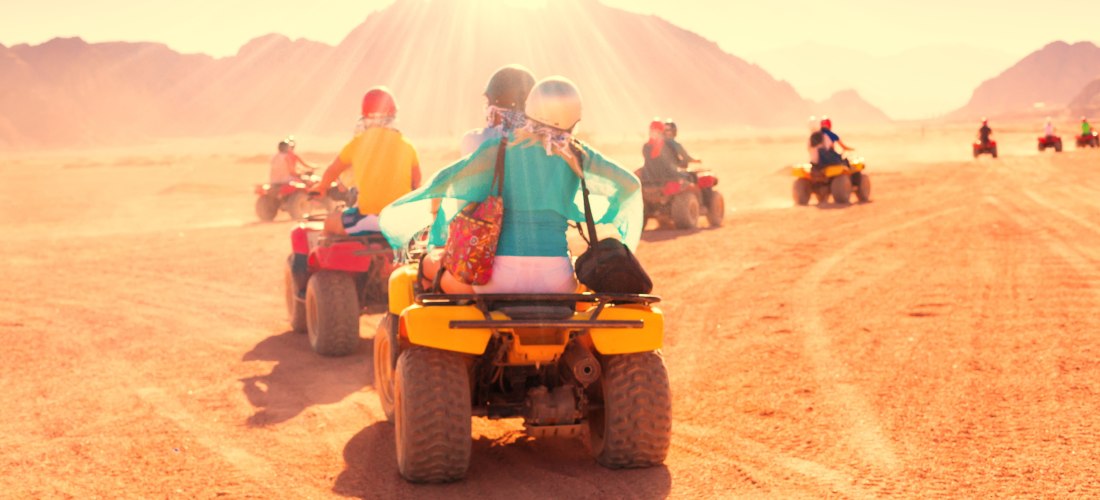 Gruppe von Menschen fährt auf Quads durch eine sonnenbeschienene Wüstenlandschaft. Im Vordergrund ein Paar auf einem gelben Quad, das bunte Kleidung und Helme trägt. Im Hintergrund sind weitere Fahrer vor einer Kulisse aus Bergen und staubigem Terrain zu 