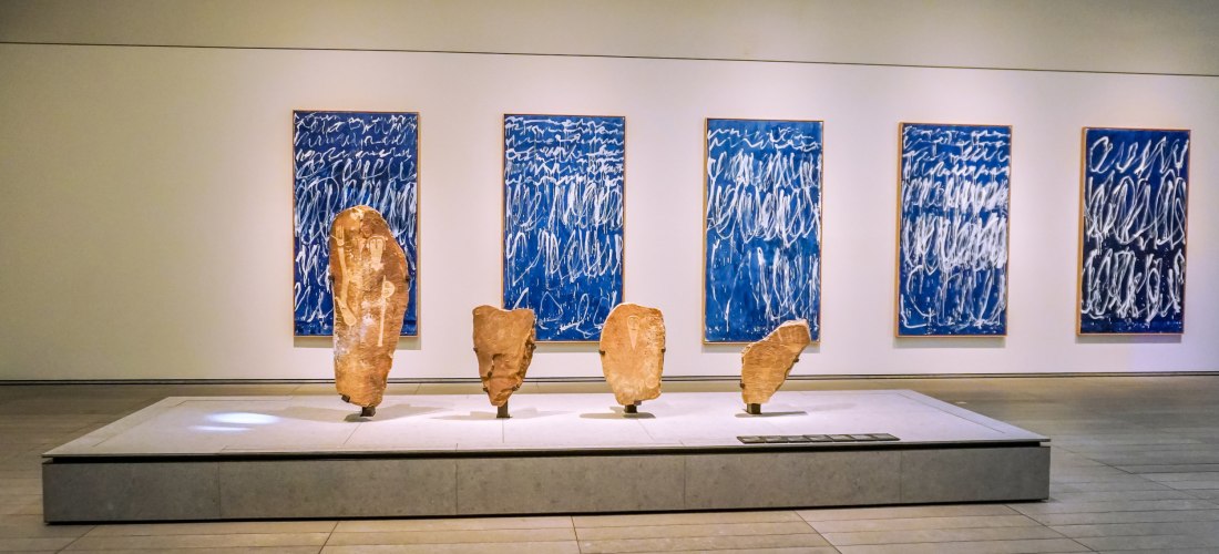 Ausstellungsraum in einem Museum mit alten Steinskulpturen auf einem Podest und fünf blauen Kunstwerken mit weißer Schrift an der Wand