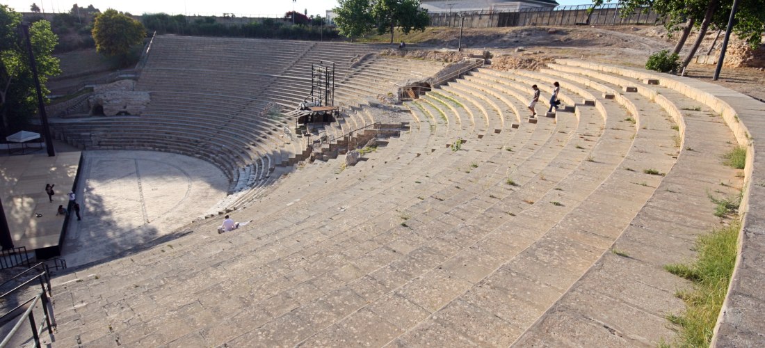 Ein römisches Amphitheater mit steinernen Sitzreihen, die sich halbkreisförmig um eine Arena legen. Im Hintergrund sind Bäume, eine moderne Moschee mit Minarett und Kuppel zu sehen. Einige Menschen sind auf den Stufen unterwegs oder sitzen dort.