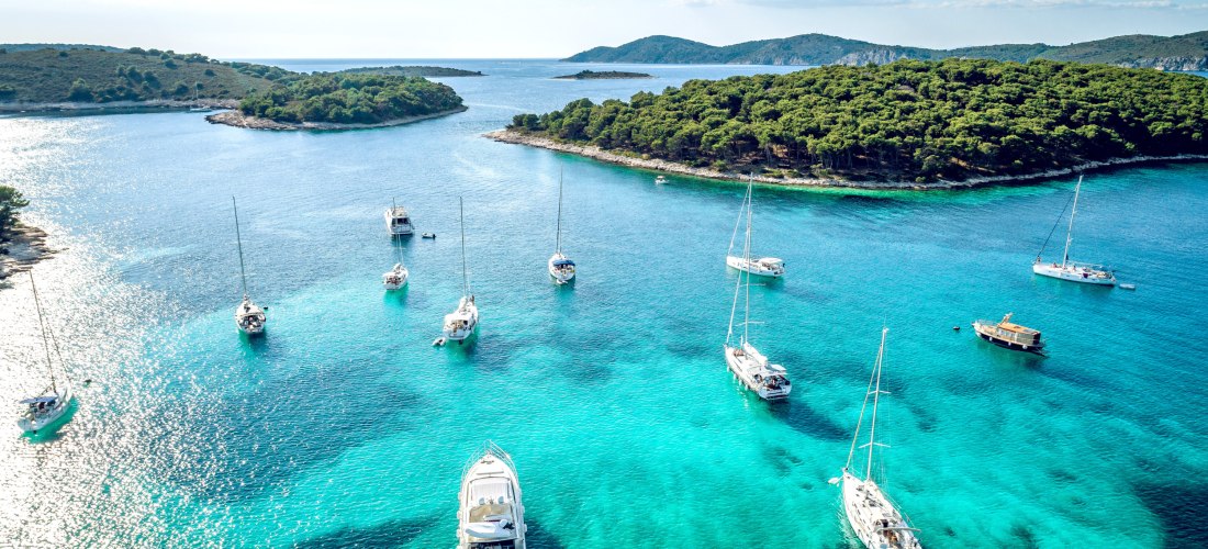 Luftaufnahme einer türkisblauen Bucht mit mehreren weißen Segelbooten und Yachten. Die Boote liegen vor einer dicht bewaldeten Insel mit sanften Hügeln im Hintergrund. Das Wasser ist klar und schimmert in verschiedenen Blautönen