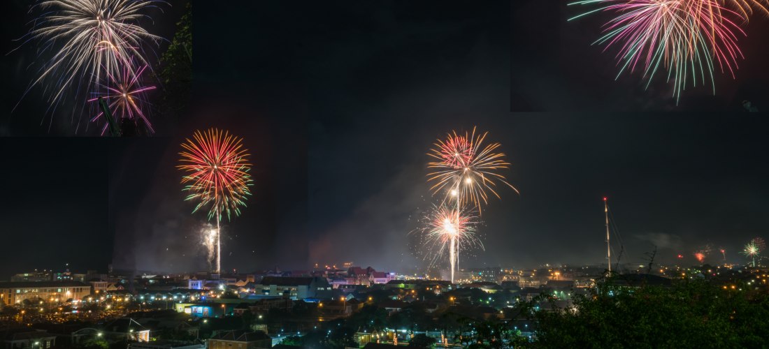 Panoramaaufnahme von Willemstad, Curaçao bei Nacht. Mehrere farbenfrohe Feuerwerke explodieren gleichzeitig über der Stadt. Die Häuser sind beleuchtet, Rauch steigt auf, und am Himmel sind rote, grüne und goldene Lichtkronen zu sehen.