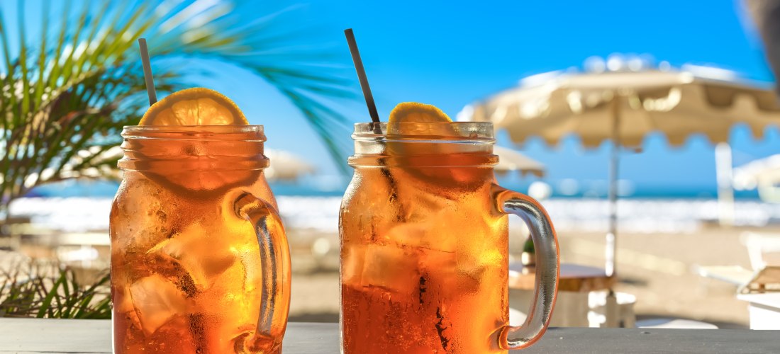Zwei Gläser mit Eistee und Zitronenscheiben auf einem Tisch am sonnigen Strand mit Palmen und Sonnenschirmen im Hintergrund