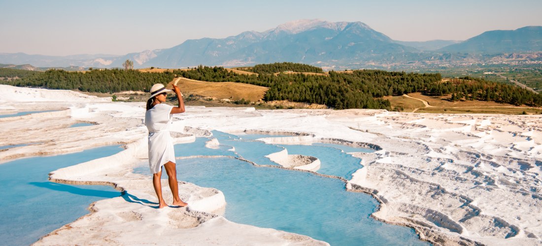 Reisende Frau steht auf den weißen Kalkterrassen von Pamukkale und blickt über die türkisfarbenen Thermalbecken mit Bergpanorama im Hintergrund