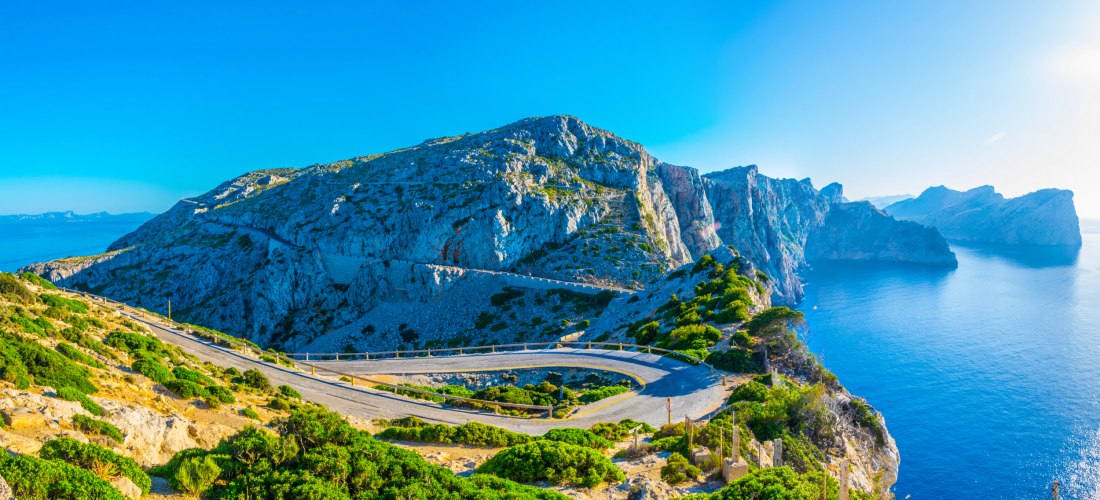 Panoramablick auf eine kurvenreiche Küstenstraße, die sich durch die felsige Berglandschaft von Cap de Formentor auf Mallorca schlängelt, mit Blick auf das tiefblaue Mittelmeer