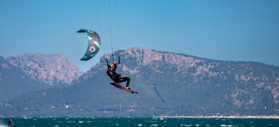 Kitesurfer in der Luft über den Wellen, mit Bergen im Hintergrund und windigem Wetter an Mallorcas Küste