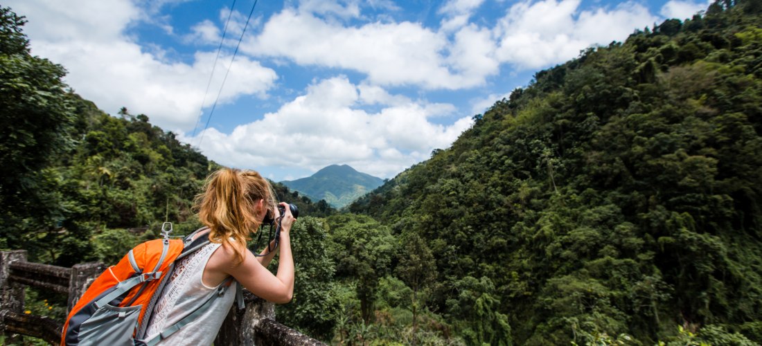 Eine junge Frau mit orangem Rucksack steht an einem Geländer und fotografiert mit einer Kamera eine üppig grüne Berglandschaft unter einem blauen Himmel mit weißen Wolken. Die Szene vermittelt ein Gefühl von Abenteuer, Natur und Freiheit.