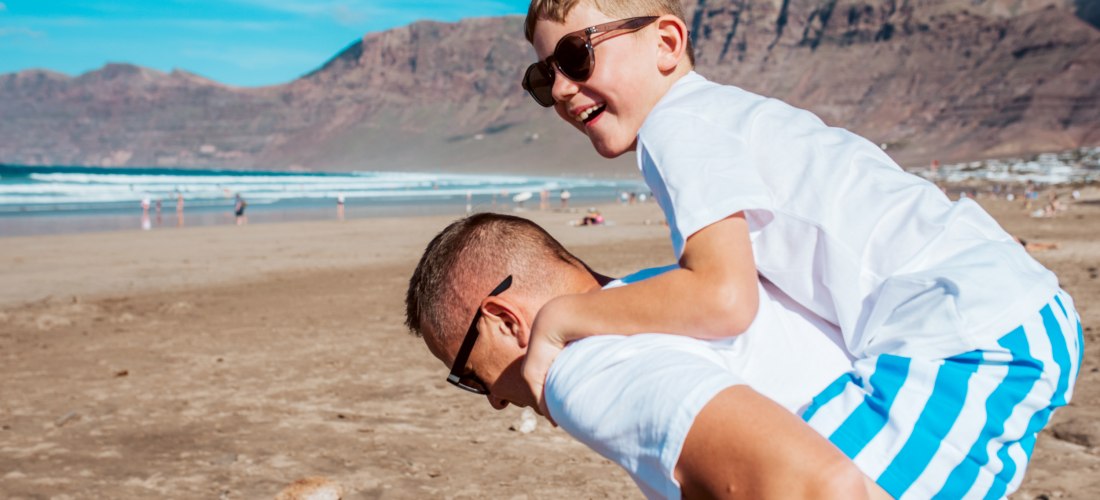Mann trägt lachenden Jungen auf dem Rücken über einen sonnigen Strand. Beide tragen Sonnenbrillen und helle Kleidung, im Hintergrund sind Meer, Berge und andere Strandbesucher zu sehen.