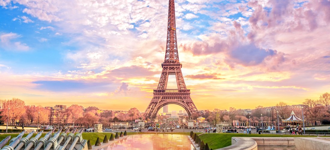 Der Eiffelturm in Paris bei Sonnenuntergang, umgeben von einem farbenprächtigen Himmel in Rosa, Orange und Blau. Im Vordergrund spiegeln sich die Farben im Wasser eines flachen Beckens. Links sind Wasserkanonen der Fontänenanlage zu sehen, rechts ein klei