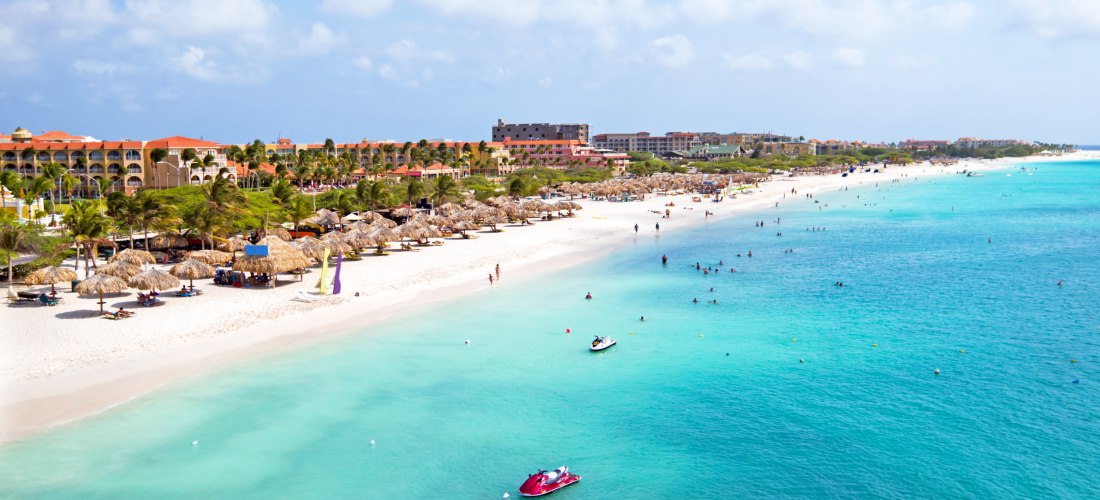 Panoramablick auf den weißen Sandstrand von Eagle Beach auf Aruba. Im türkisblauen Wasser sind Jet-Skis unterwegs, während zahlreiche Menschen im Wasser baden oder am Ufer spazieren. Am Strand stehen Palapa-Sonnenschirme und Liegen. Dahinter reihen sich R