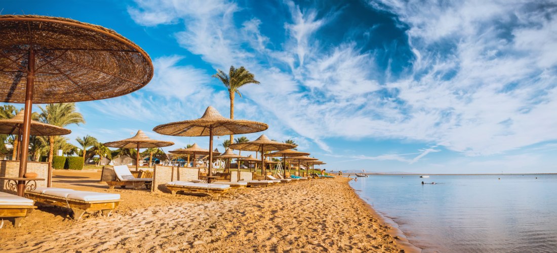 Sonnenverwöhnter Sandstrand mit zahlreichen Sonnenliegen und Sonnenschirmen aus Naturmaterialien. Palmen säumen den Hintergrund, der ruhige Ozean erstreckt sich unter einem teils bewölkten, blauen Himmel. Einige Menschen baden im Wasser oder entspannen am