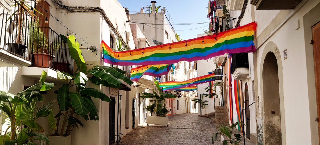 Eine schmale Gasse in Ibiza-Stadt, gesäumt von weiß gestrichenen Häusern mit Balkonen und Pflanzen. Mehrere große Regenbogenflaggen hängen quer über die Straße, als festlicher Schmuck. Die Szene ist sonnig und friedlich.