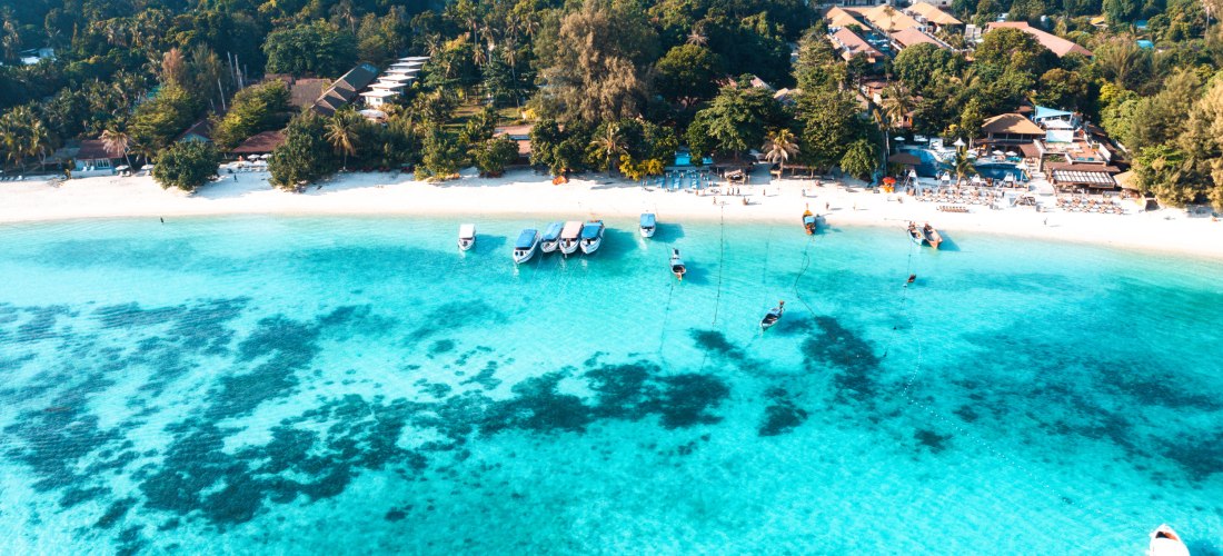 Luftaufnahme eines tropischen Resorts mit weißen Sandstränden und türkisblauem Wasser. Mehrere Boote ankern vor der Küste, dahinter dichte grüne Vegetation und Hotelanlagen.